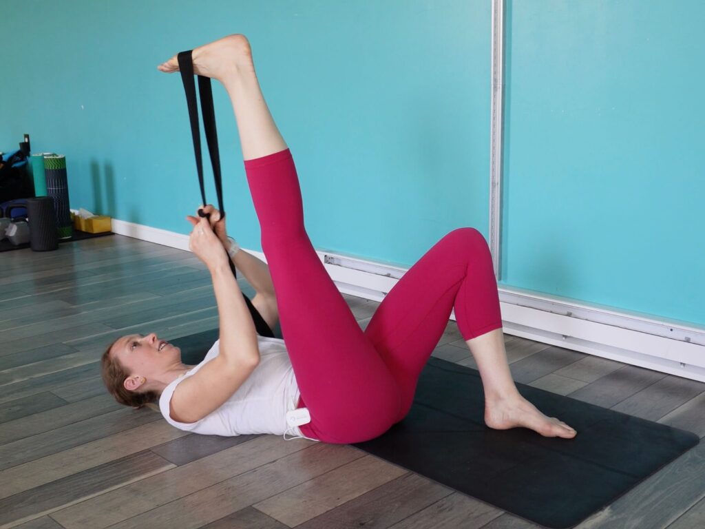 Dr. Chloe laying on her back with one leg straight up in the air and the other with his foot on the ground. She is using a strap around her toes of the extended leg to stretch the hamstring